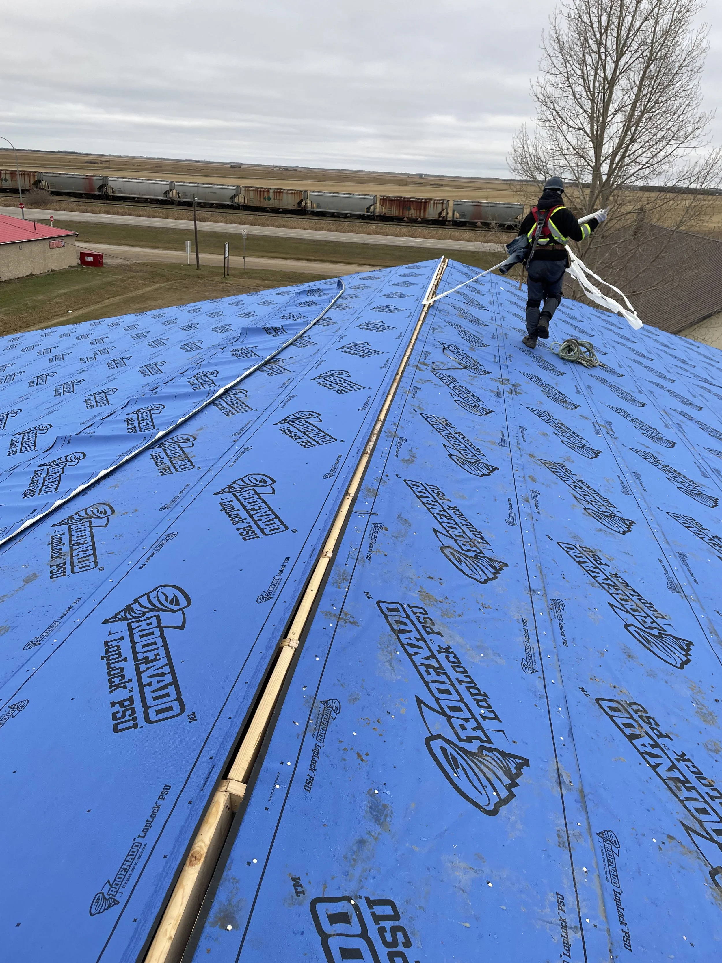 A person working on a roof with blue underlayment, wearing protective clothing and a helmet, near a tree and train tracks in a rural area.
