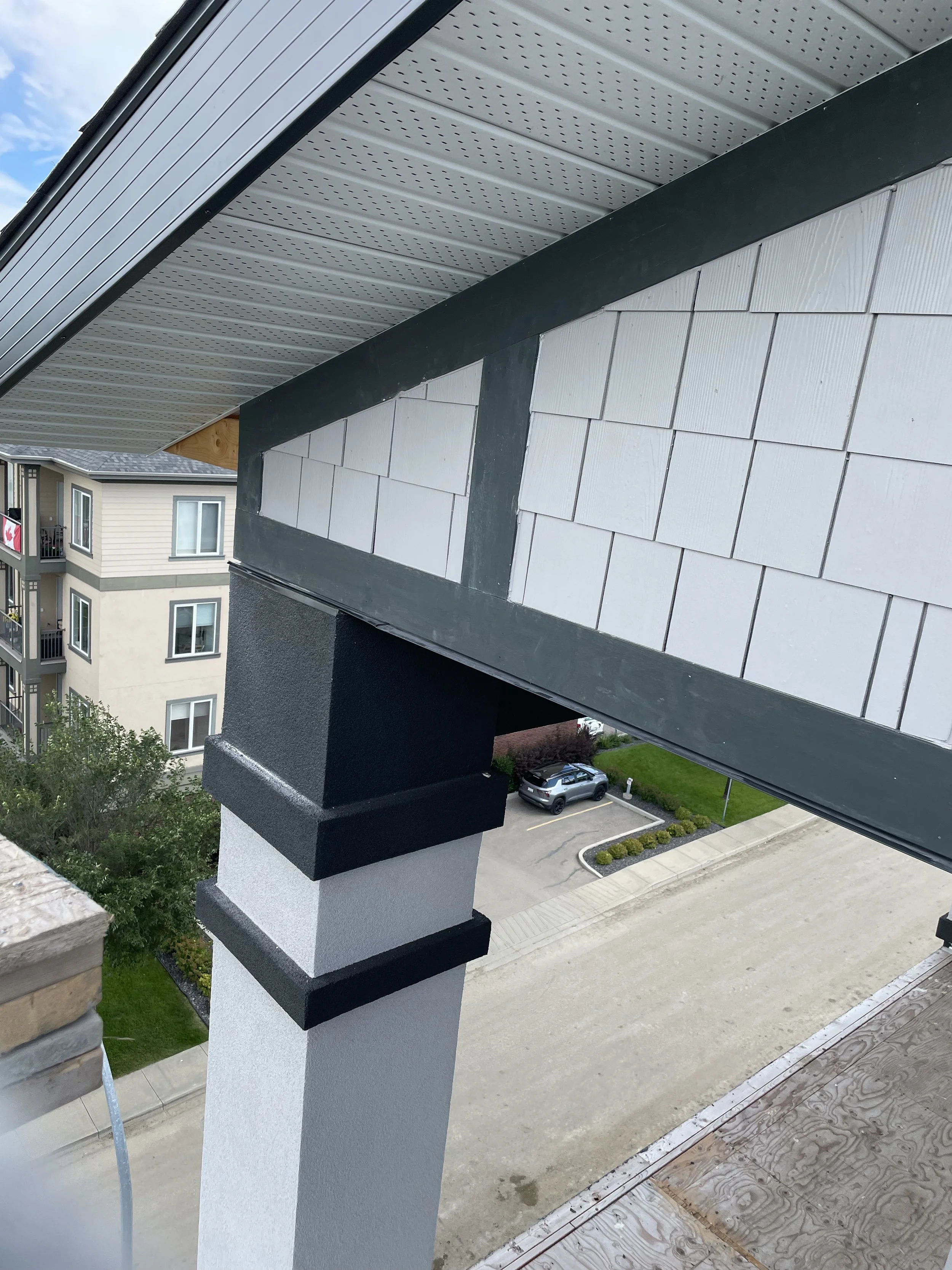 Close-up of a building's exterior corner showing white siding with black trim, a black and white column, and part of the roof with metal soffit panels. In the background, a parking lot with cars and another apartment building are visible.