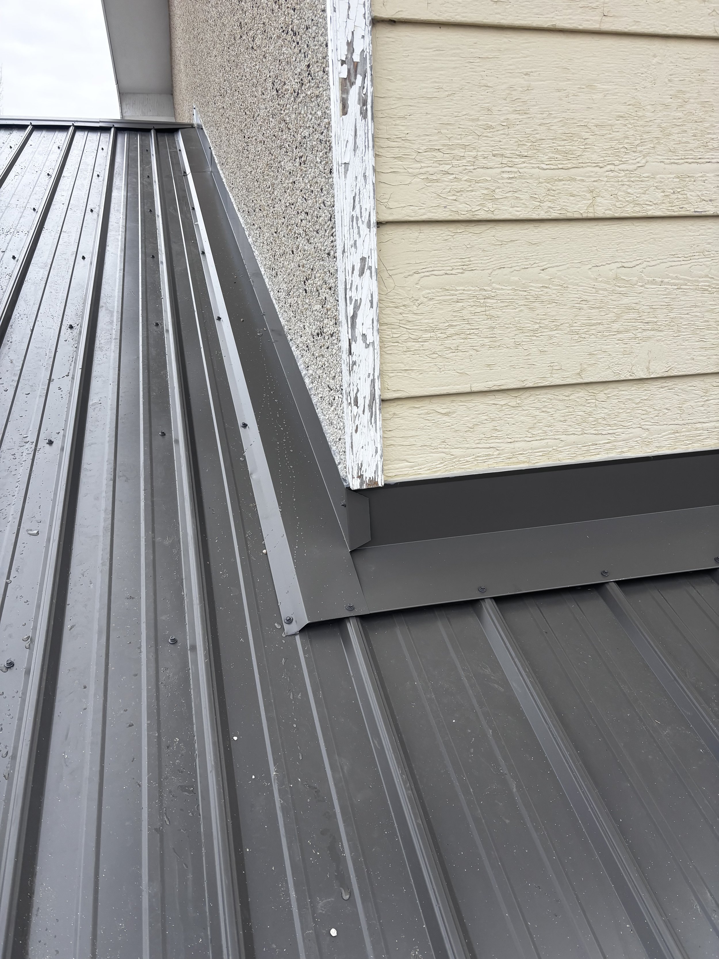Close-up of a metal roofing on a building, showing a gray metal seam with water droplets, and a corner where the roof meets a beige and off-white exterior wall with textured siding.