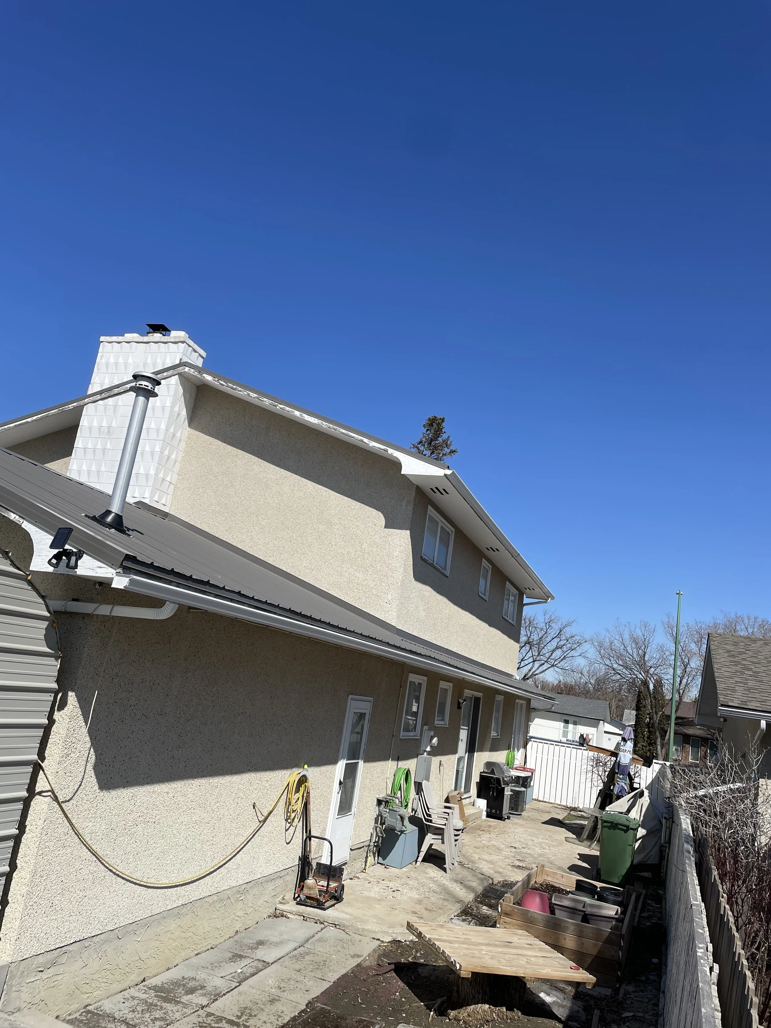 Backyard of a two-story beige house with a gray roof, with various outdoor items, garden hose, chairs, trash bins, and a white fence, under a clear blue sky.