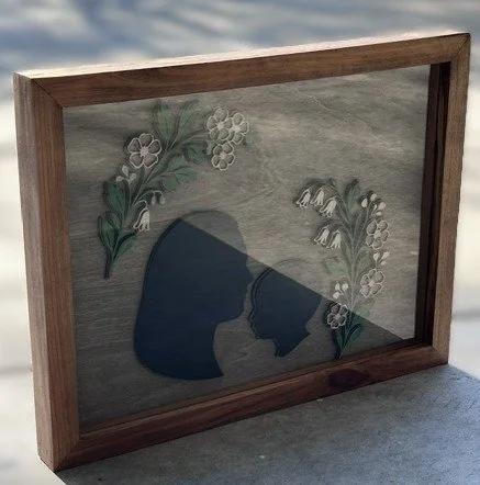 A wooden-framed shadow box with a painted background of flowers and leaves, and a silhouette of a woman and child touching foreheads.