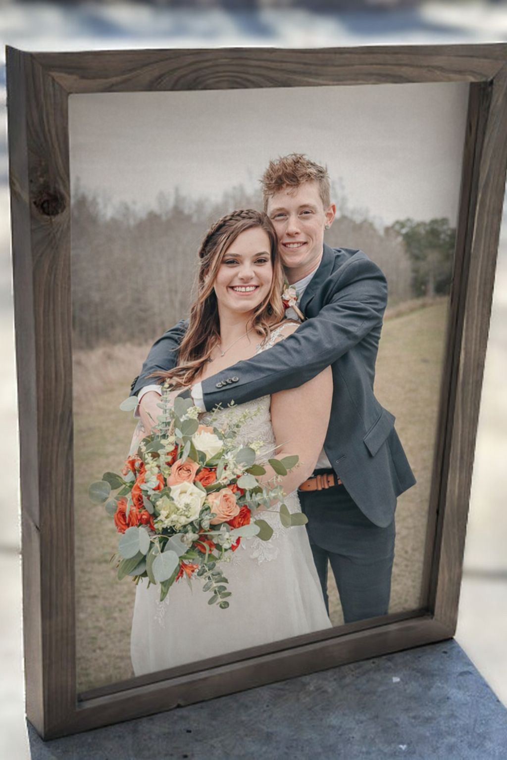 A framed photograph of a smiling bride and groom outdoors, with the groom hugging the bride from behind. The bride holds a bouquet of white, red, and orange flowers with greenery. The background shows a grassy field and trees.
