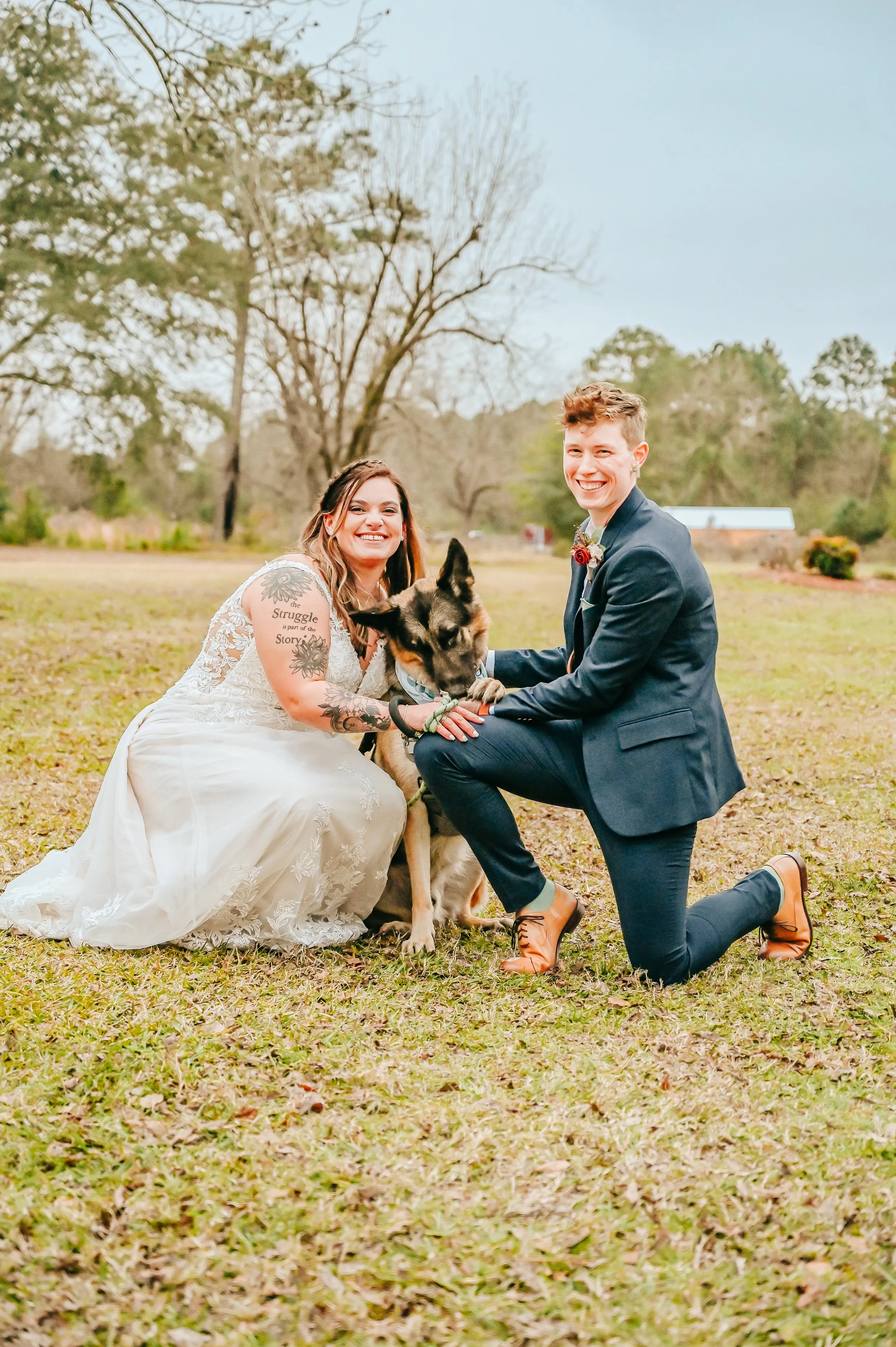 A couple dressed in wedding attire kneels on a grassy field with their dog, smiling at the camera with the woman wearing a white wedding dress and the man in a dark suit.
