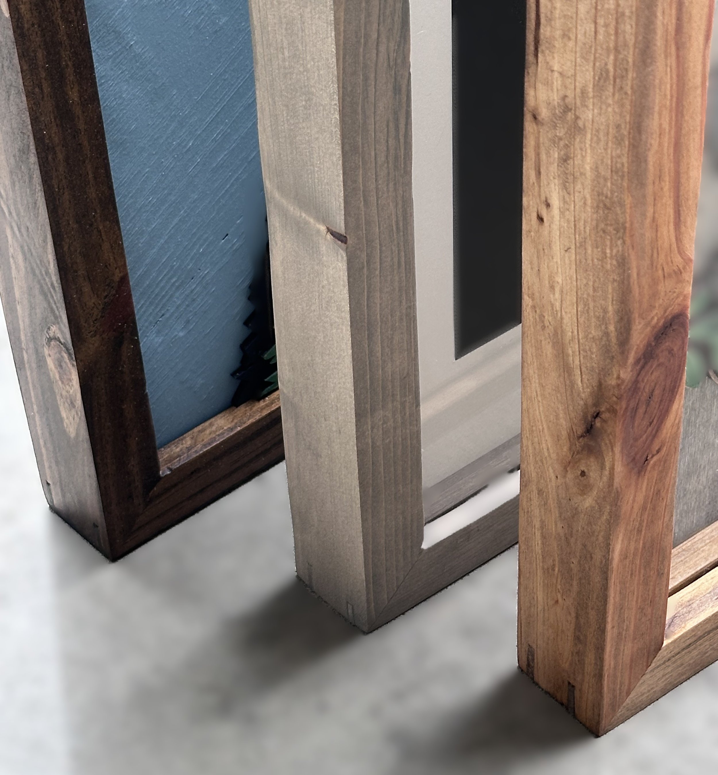 Close-up of three wooden picture frames in different colors and finishes leaning against a gray surface.