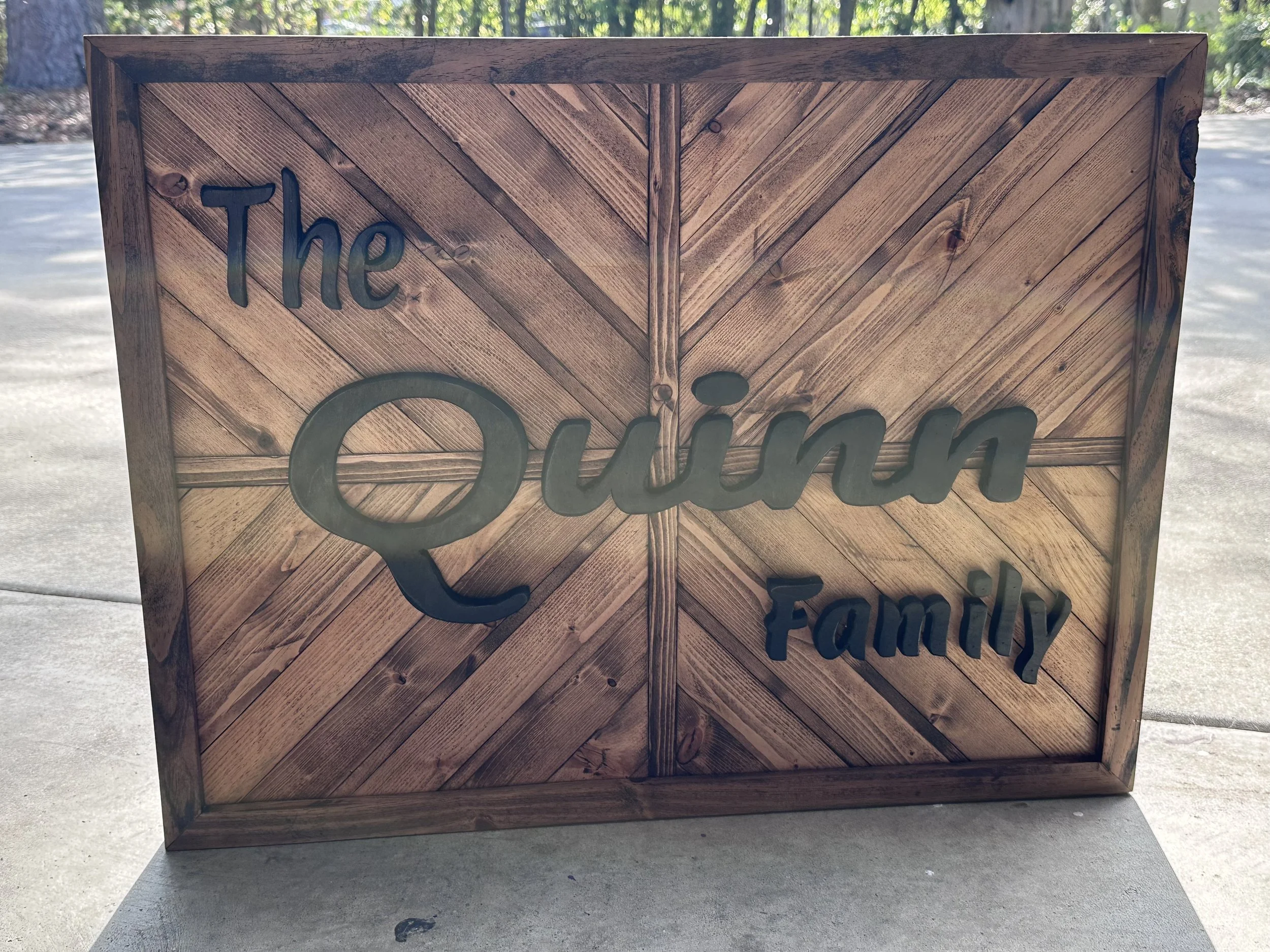 Wooden sign with the words "The Quin Family" in black, decorative script, set against a light, chevron-patterned wooden background, outdoors on concrete.