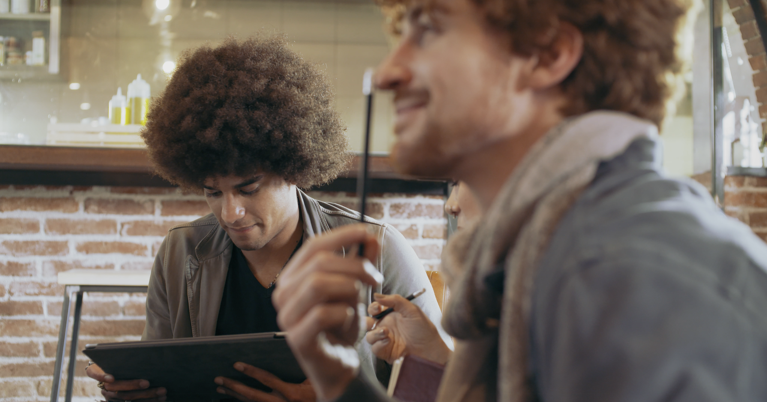 Two young men with curly hair, one with an afro and the other with a chestnut-colored afro, sitting at a table in a cozy, rustic cafe with a brick wall background. They are engaged in looking at a menu or tablet, smiling and sharing a moment of conversation.