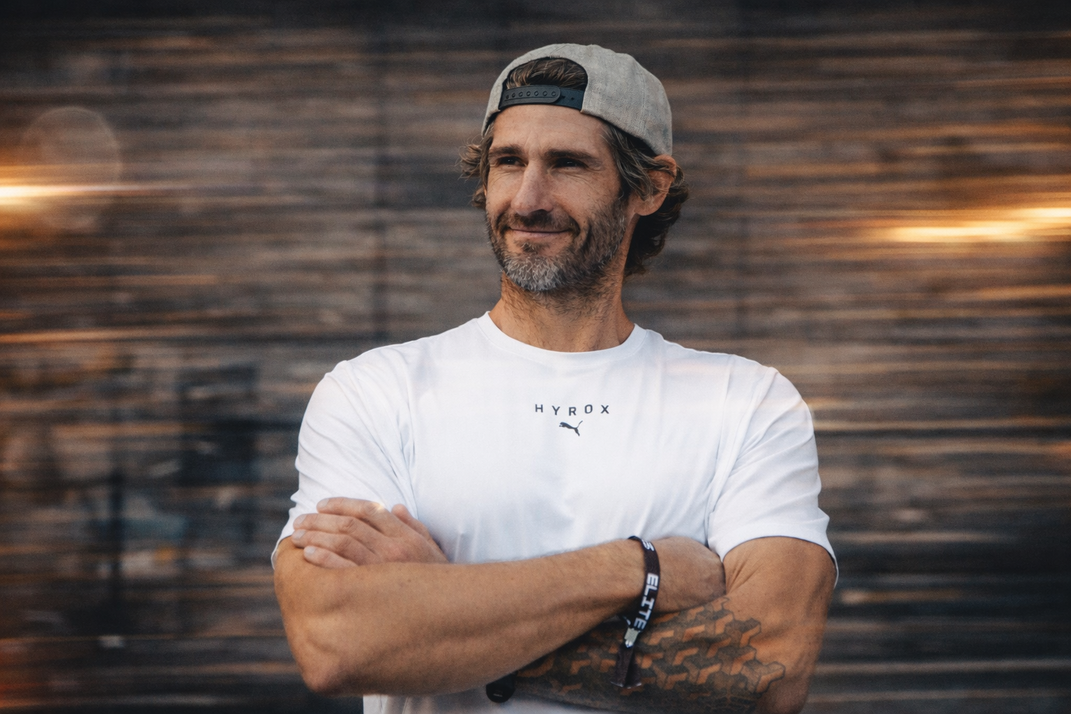 A man with gray and brown hair, beard, and light skin, wearing a gray backward baseball cap and a white t-shirt with the Puma logo, standing with arms crossed against a blurred wooden wall background.