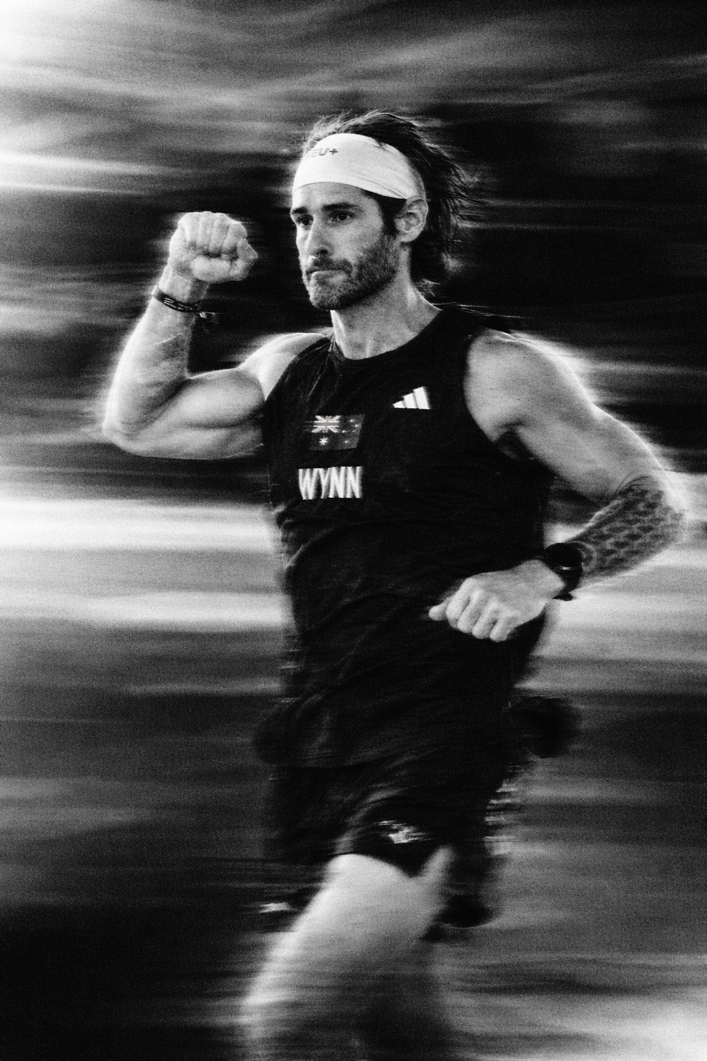 A man running outdoors in black athletic clothing, wearing a headband, watching a fitness tracker or watch, with a tattooed arm, and the background is blurred to suggest motion.