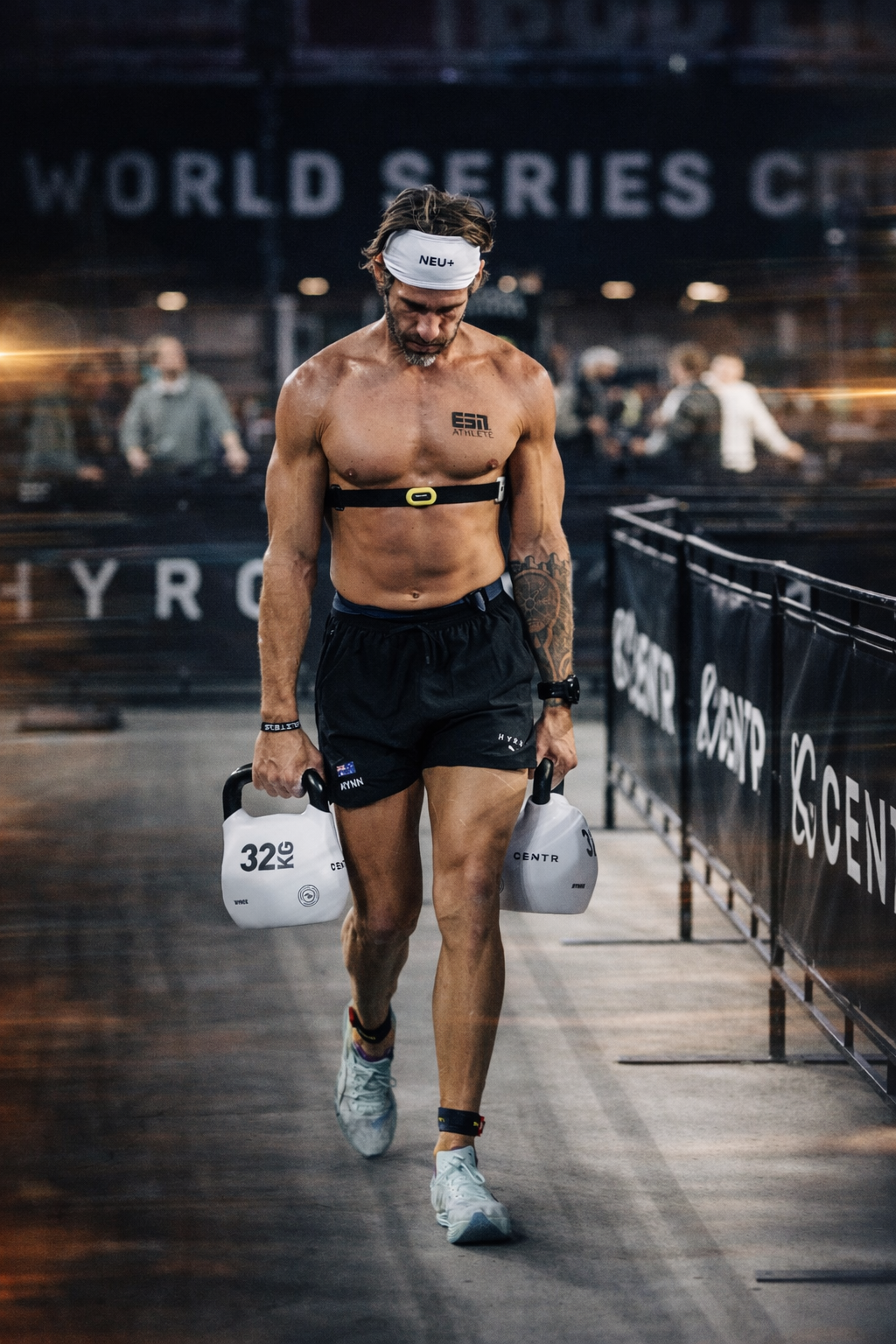 A shirtless male athlete with a headband, carrying two 32 kg kettlebells, walking through a gym with