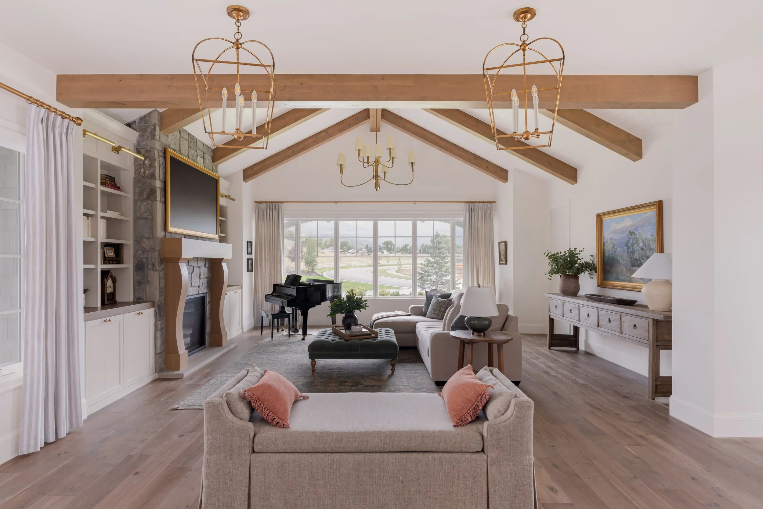 Spacious living room with vaulted ceiling, wooden beams, large window, beige sofa, armchair, green ottoman, potted plant, fireplace, TV, and a chessboard on a side table.