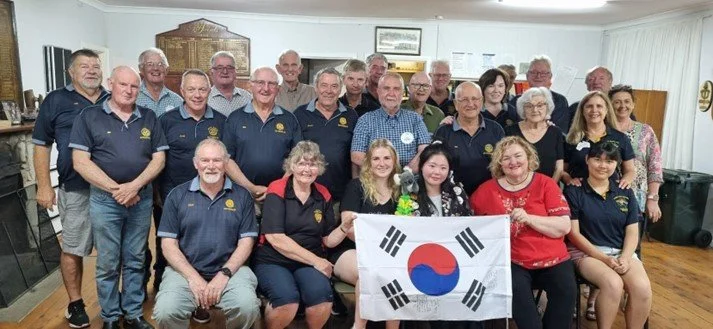 A large group of people, mostly older adults, gathered indoors for a group photo. Some are wearing matching navy blue shirts with a logo, and others are in casual attire. They are holding a South Korean flag. The room has white curtains, and there ar