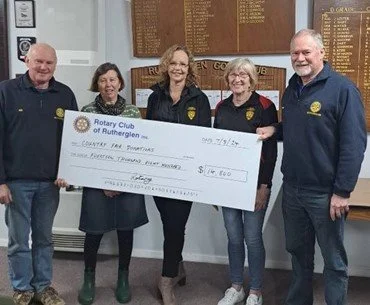 Five people standing together holding a large check with a Rotary Club banner, in an indoor setting.