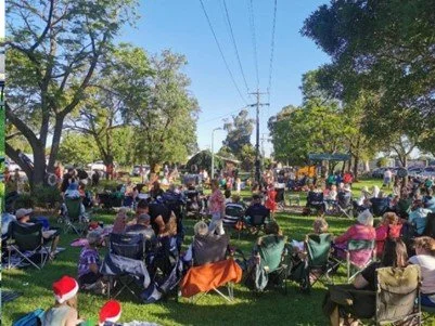 A large outdoor gathering in a park with people seated on chairs and blankets, some wearing Santa hats, with trees and utility poles visible in the background.