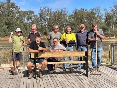 A group of seven people outdoors on a wooden deck, with trees and blue sky in the background. They are posing around a picnic table, with some standing and one person sitting, smiling at the camera.
