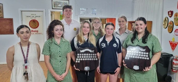 Group of seven students in school uniforms standing in a classroom, with two holding award plaques, smiling at the camera.