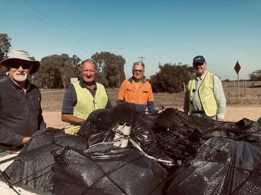 Four men standing outdoors behind a large pile of black bags, with trees and power lines in the background.