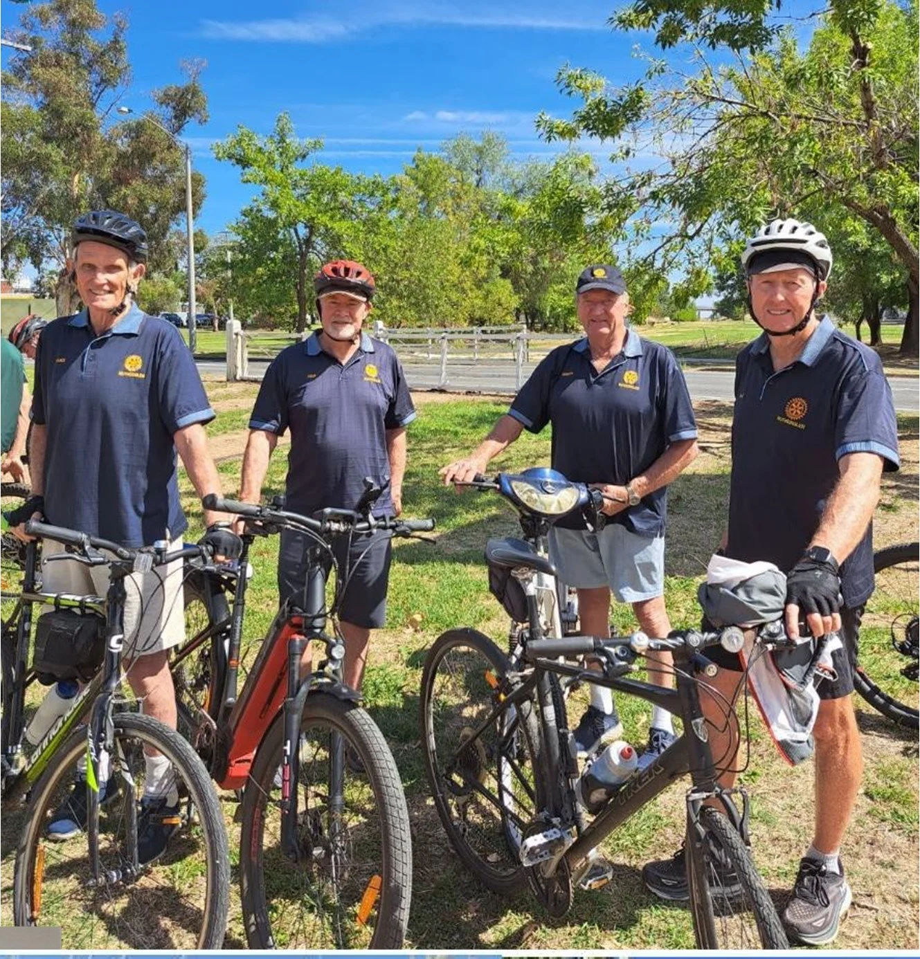 Four older men standing outdoors with their bicycles on a sunny day, wearing helmets and matching navy shirts, smiling at the camera.