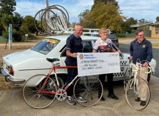 Three men standing outdoors next to a vintage white car with bicycles in front, holding a large check, with a bike sculpture and trees in the background.