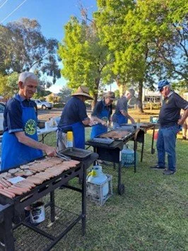 People grilling sausages outdoors in a park with trees and blue sky.