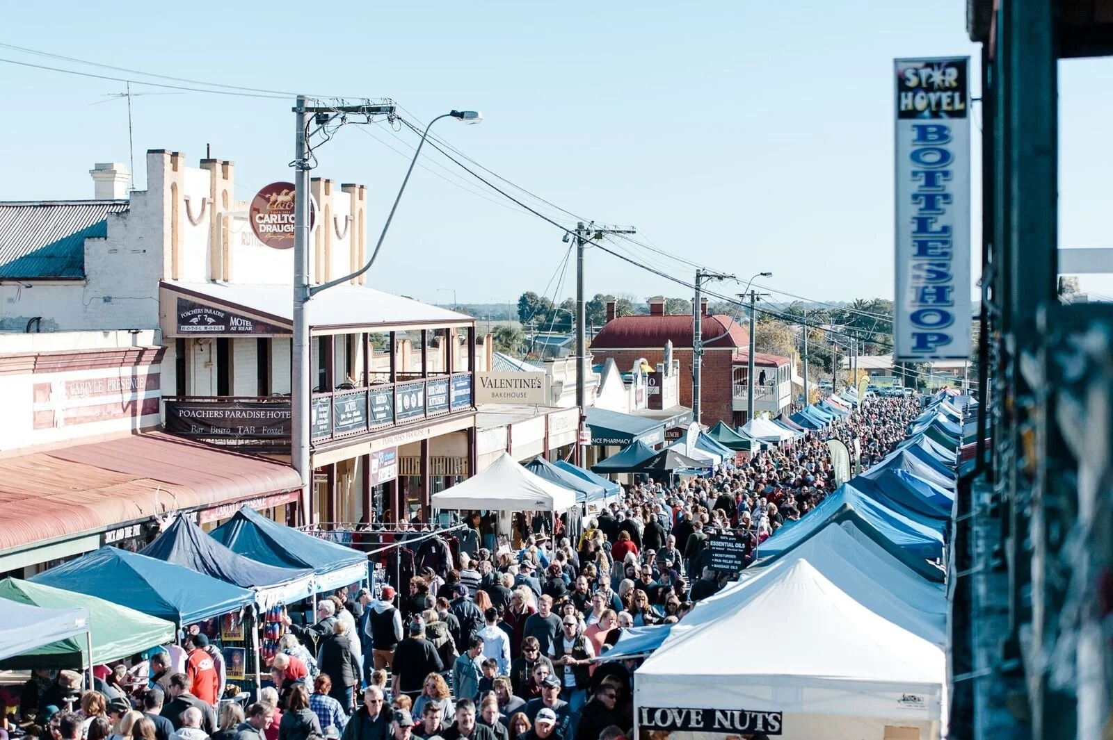 Crowd walking along a street with market stalls and tents, buildings with signs including Motel and Bakehouse, and clear blue sky.