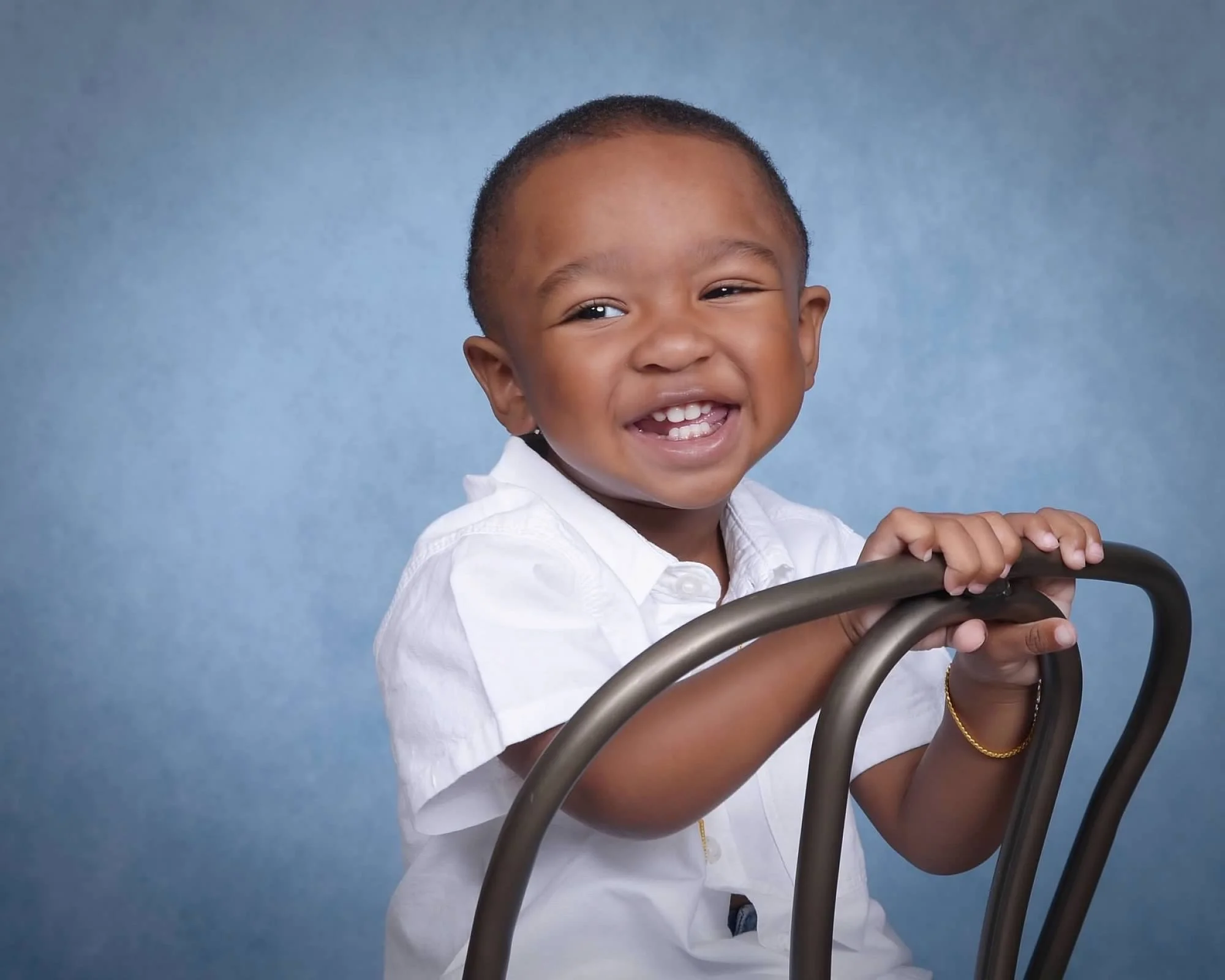 A toddler smiling, wearing a white shirt, siting on a metal chair backwards with a blue background.