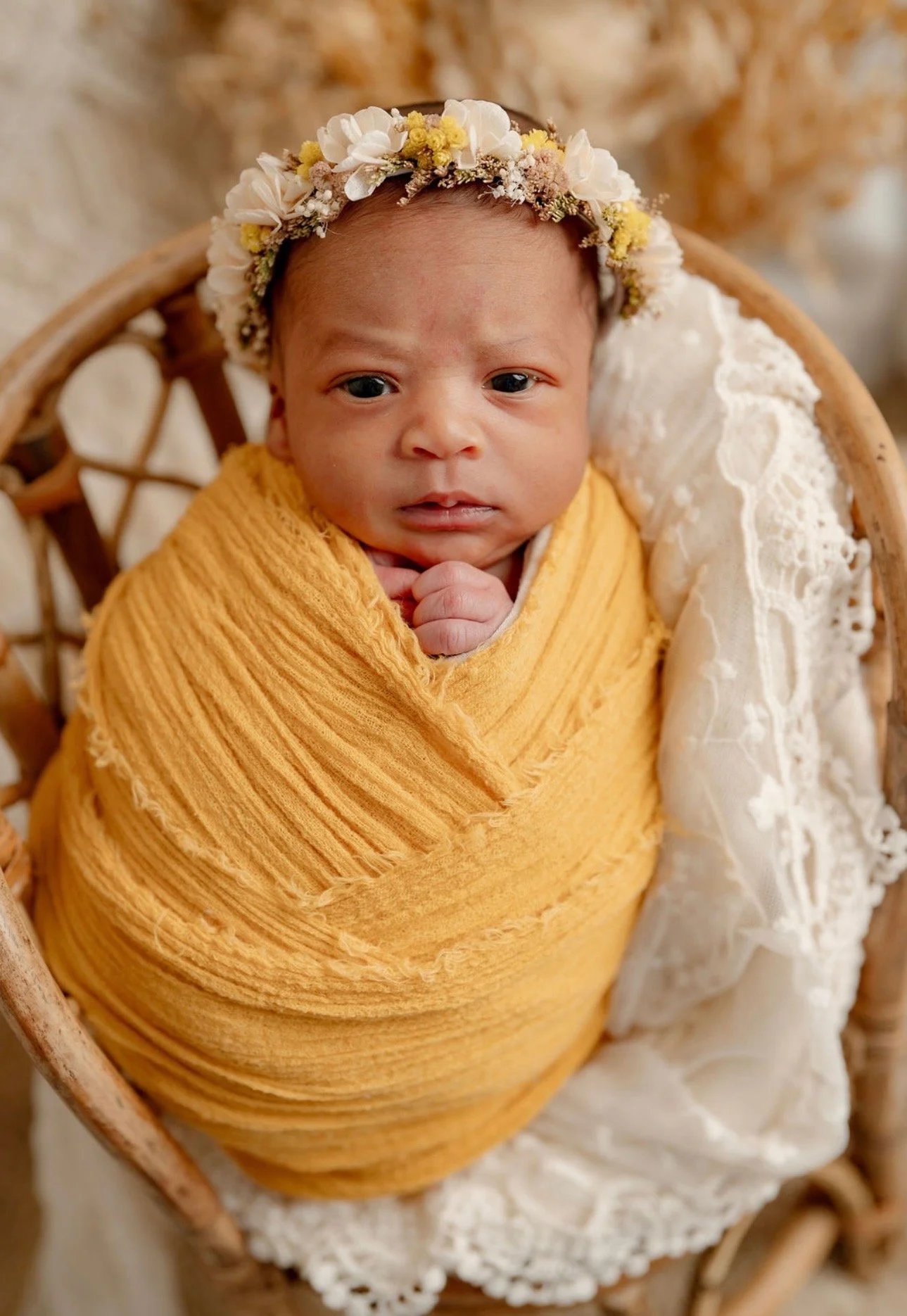 A baby wrapped in a mustard yellow blanket, lying in a wicker basket, wearing a flower crown with white and yellow flowers, and a white lace outfit.