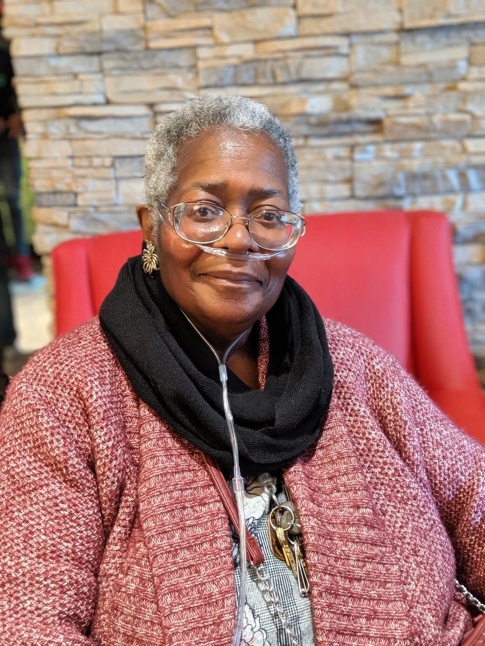 An elderly woman with gray hair, wearing glasses and a nasal oxygen tube, sitting on a red chair in front of a stone wall, dressed in a pink textured jacket and a black scarf.