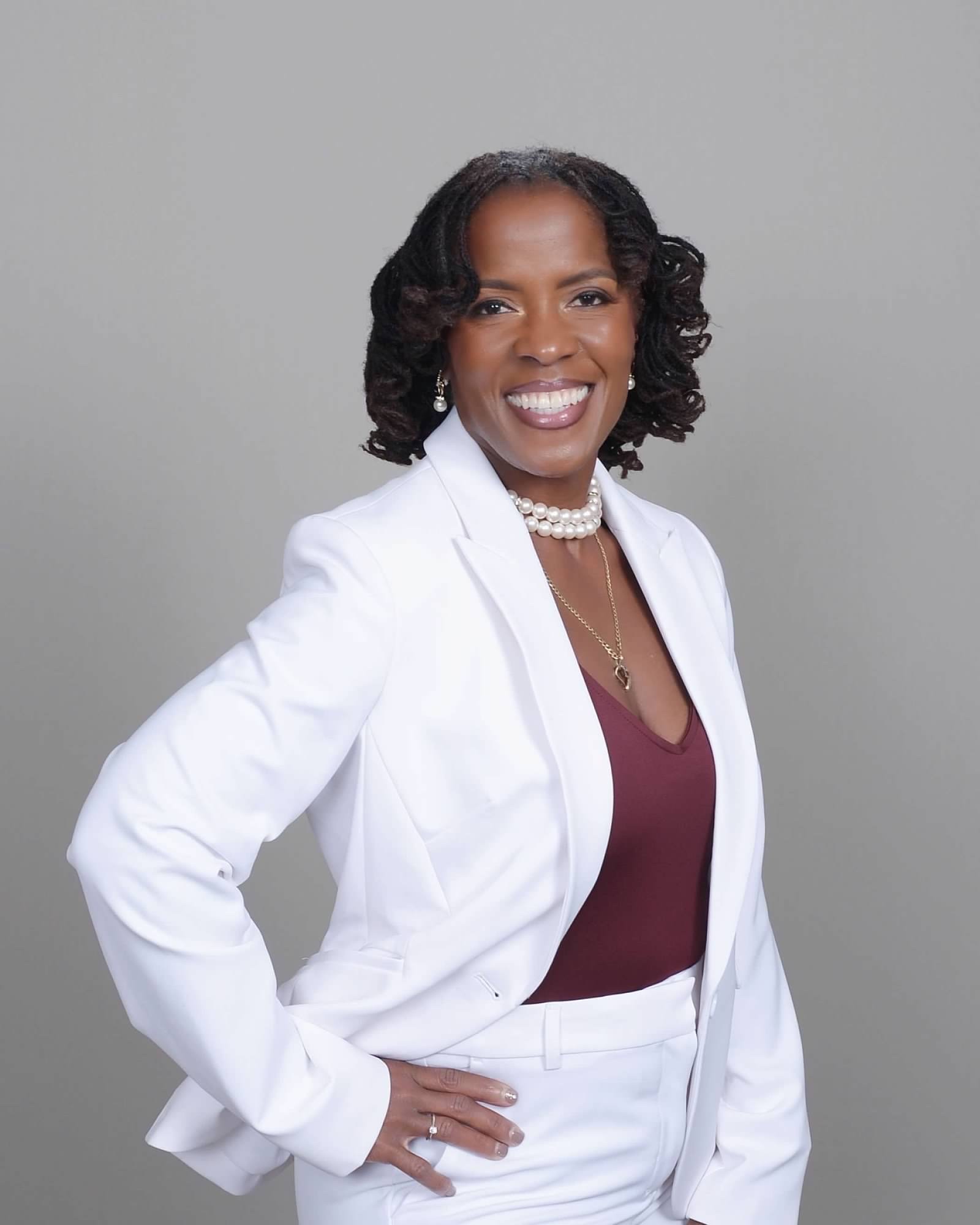 A smiling woman with dark, styled hair, wearing pearl earrings, a pearl necklace, and a white blazer over a maroon top, standing against a plain gray background.