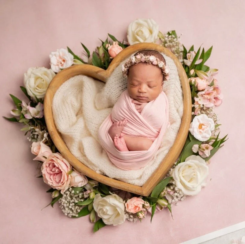 A newborn baby girl wrapped in pink blankets, sleeping peacefully in a wooden heart-shaped bed, surrounded by pink and white flowers on a pink background.