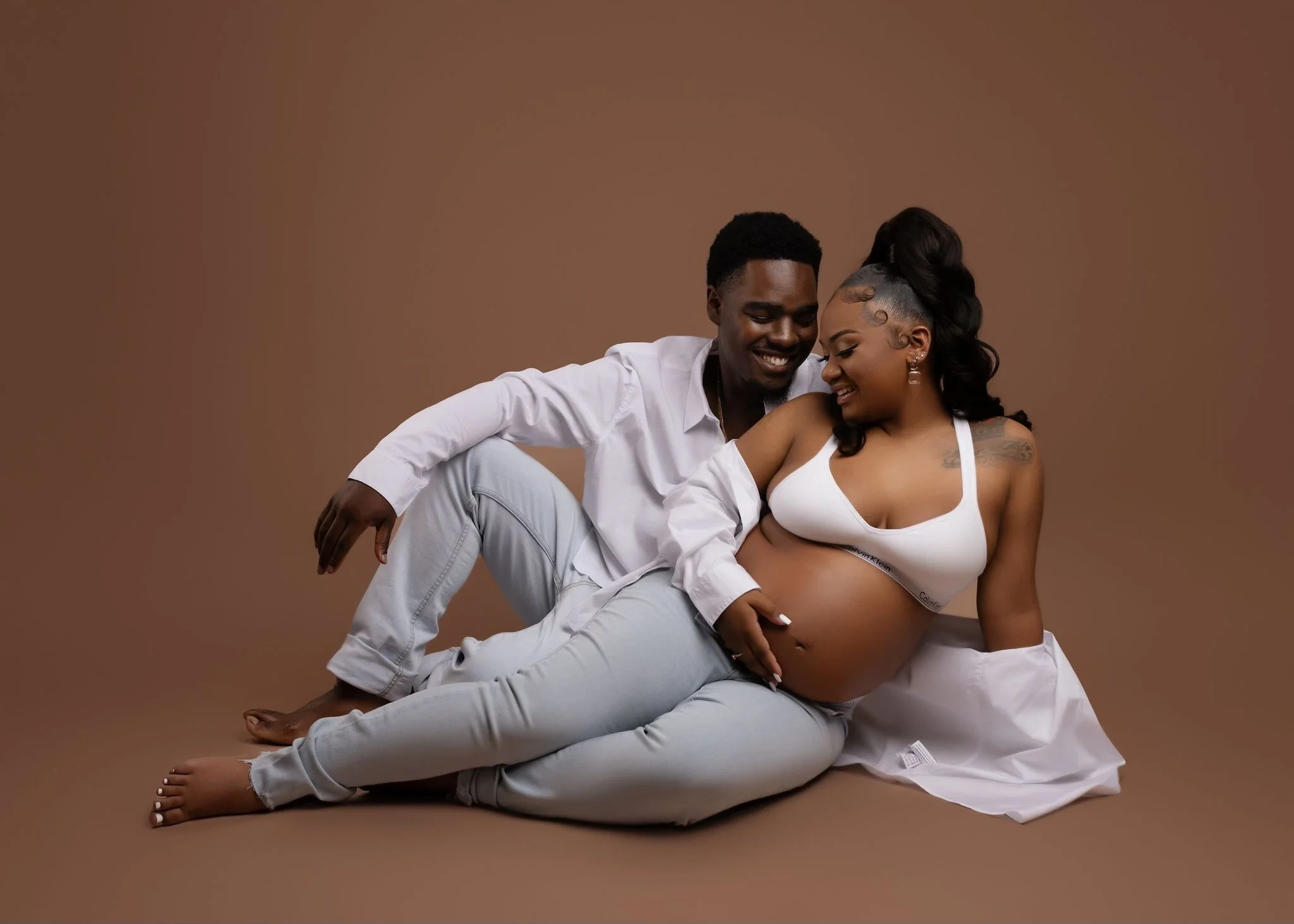 A couple sitting on the floor with a pregnant woman wearing a white crop top and white pants, and a man in a white shirt and light gray pants, smiling and looking at each other against a brown background.