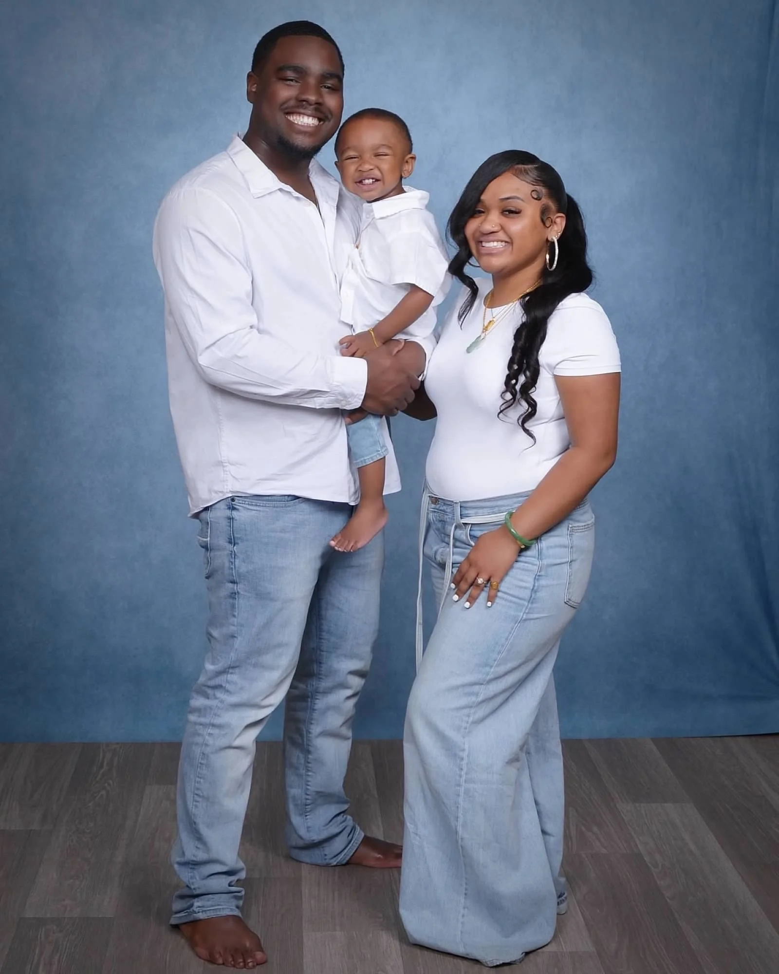 A family of three posing together against a blue backdrop. The man on the left is smiling, holding a smiling toddler in his arms, who is wearing a white shirt. The woman on the right, also smiling, is dressed in a white shirt and light blue jeans, with long black hair styled in side curls.