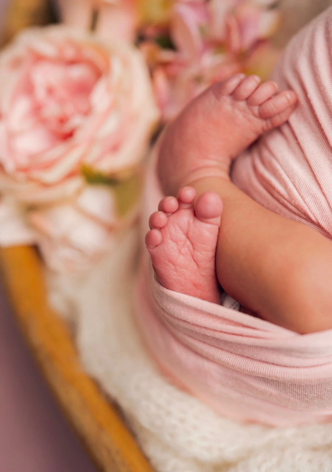 Close-up of a newborn baby's hand resting on their chest, wrapped in pink fabric, with blurred pink roses in the background.
