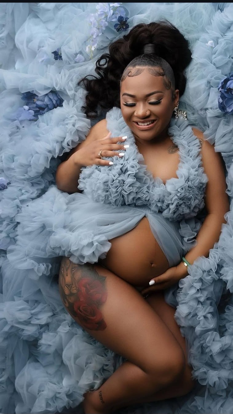 A pregnant woman is lying on a bed surrounded by blue and white ruffled fabric and flowers. She has long dark curly hair, makeup, and is smiling. She is wearing a ruffled blue top, has a tattoo of roses on her thigh, and is holding her belly with one hand while the other is on her chest.