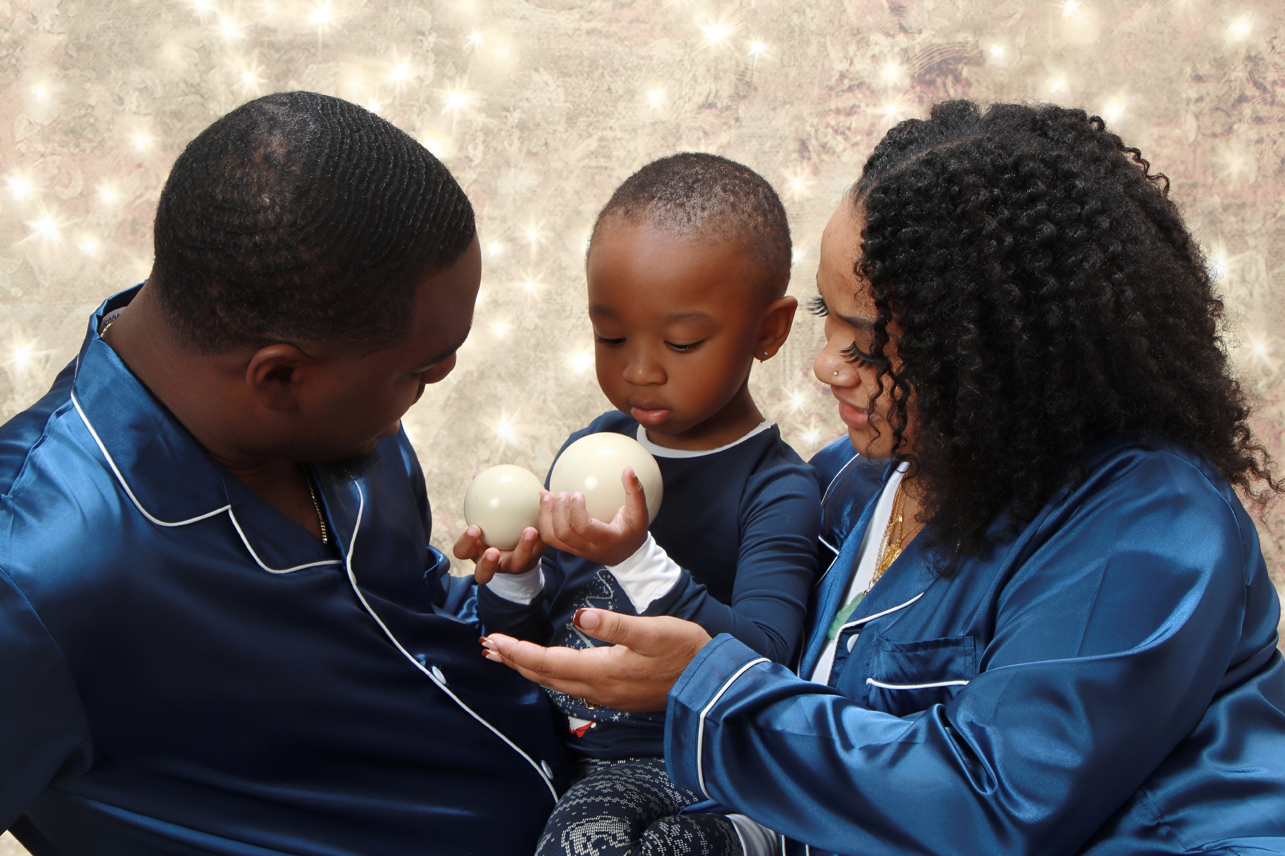 Family of three, a man, woman, and young girl, dressed in matching blue satin pajamas, holding and examining three white Christmas ornaments together, standing in front of a sparkly background.
