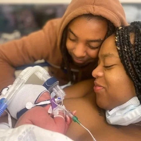 A young couple lovingly touching foreheads while holding a newborn baby with medical tubes and a hat, in a hospital setting.