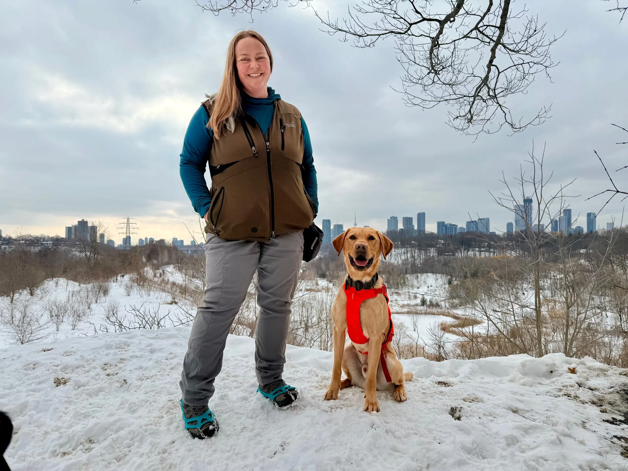 A woman and a dog on a snowy hill with a city skyline in the background. The woman is smiling, wearing a brown vest, blue long-sleeve shirt, gray pants, and hiking shoes. The dog is a golden retriever mix, sitting, wearing a red harness and looking at the camera.