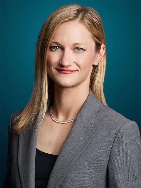 Professional woman with blonde hair and blue eyes, wearing a gray blazer and a pearl necklace, smiling against a teal background.