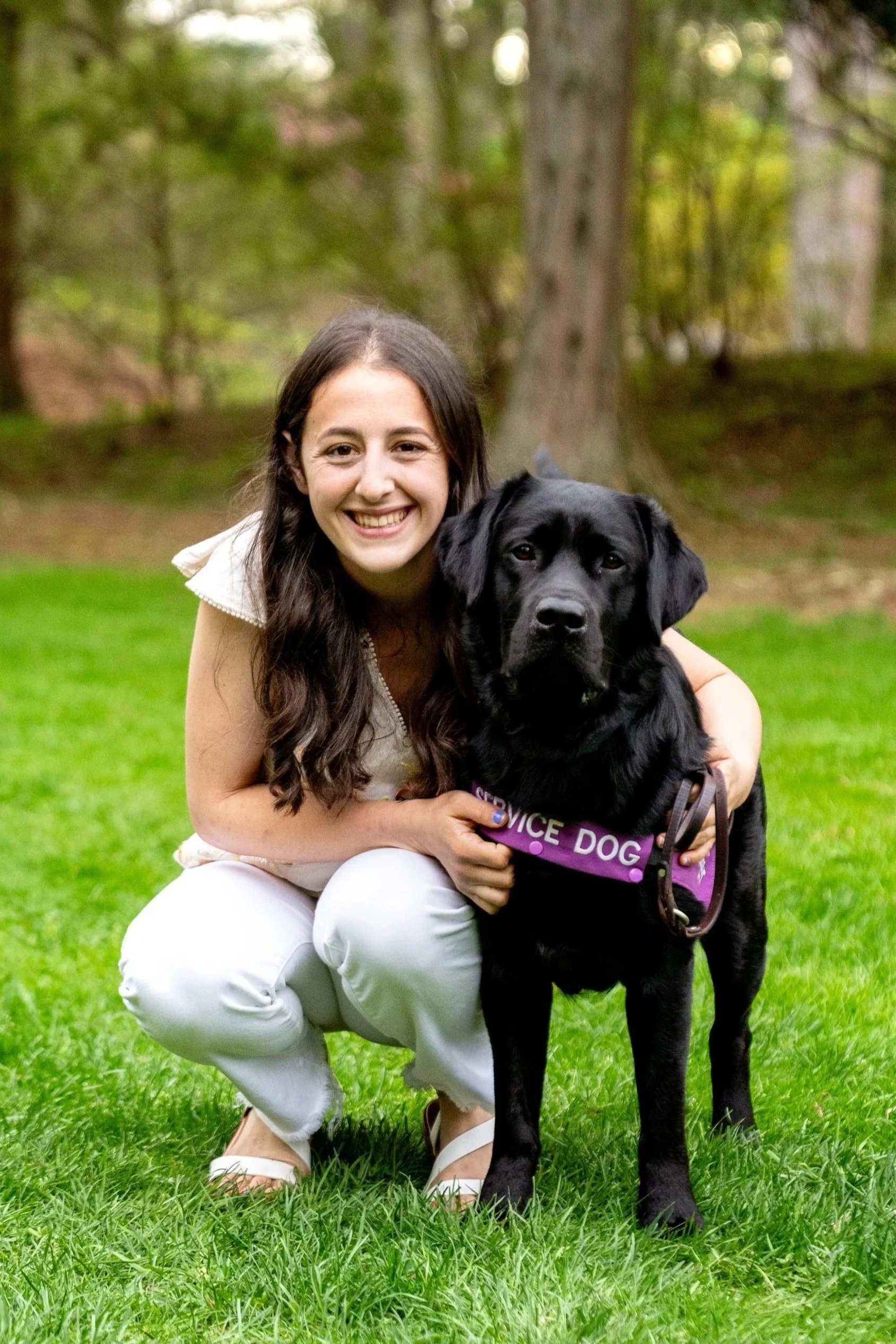 A smiling woman squatting on green grass, hugging a black service dog wearing a purple vest that says "SERVICE DOG". They are outdoors with trees in the background.