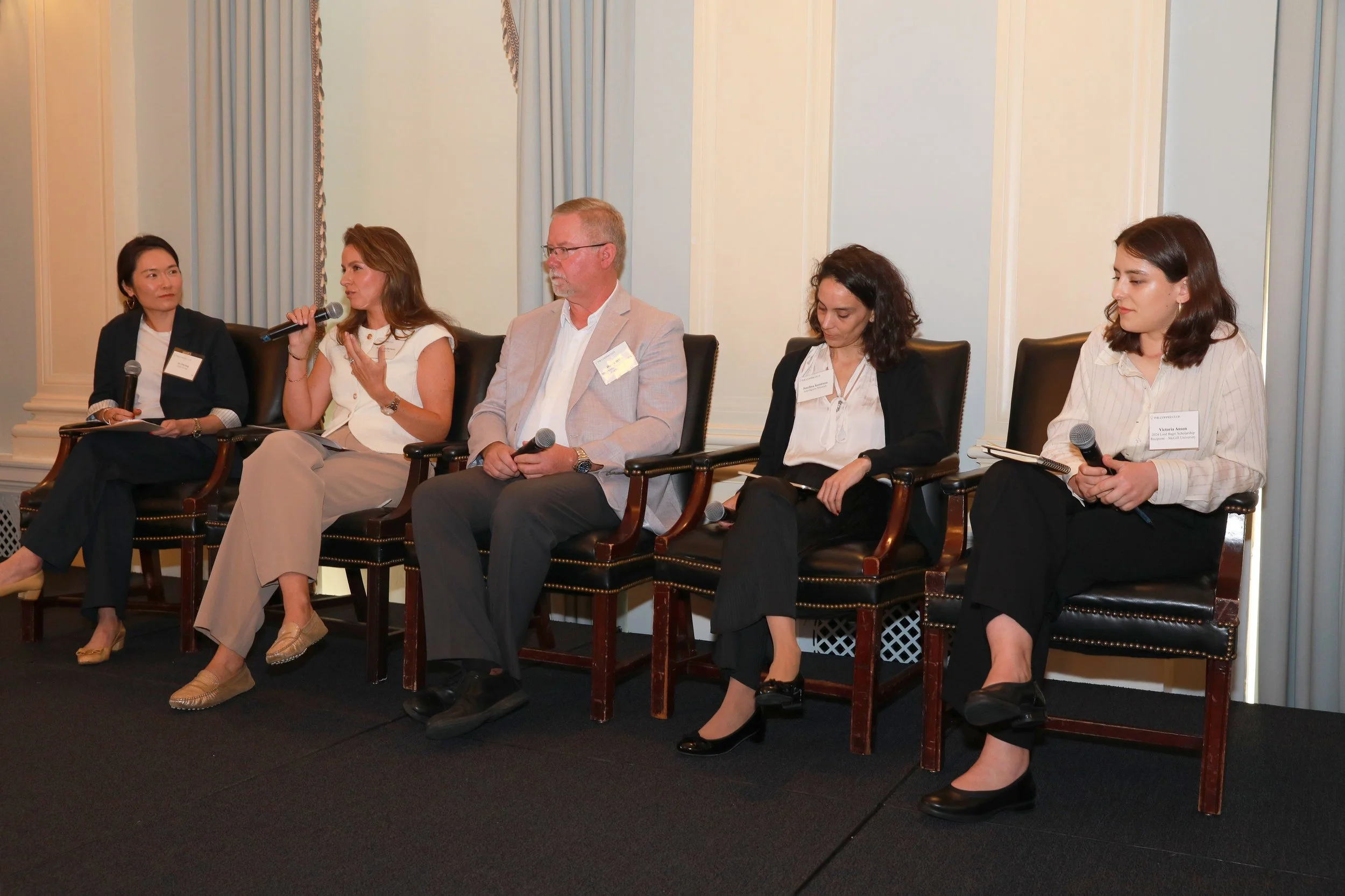 Five people sitting on wooden chairs on a stage, participating in a panel discussion. All hold microphones, and they are dressed in business casual attire. The background features light-colored walls with curtains.