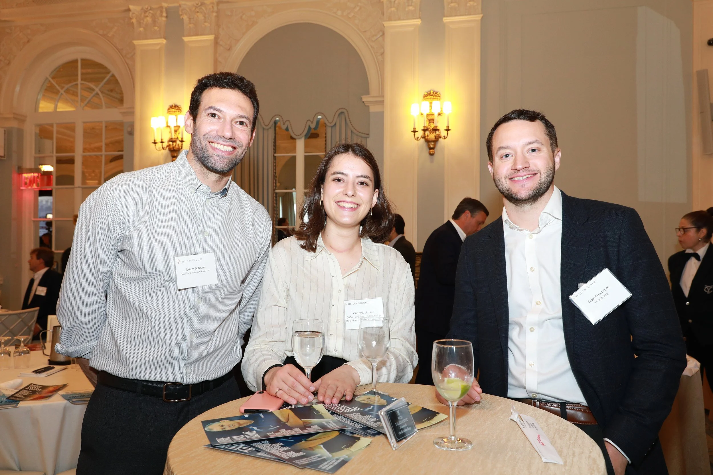 Three smiling people stand around a round table at a formal event, with a man on each side of a woman. The room has ornate decor and bright lighting.