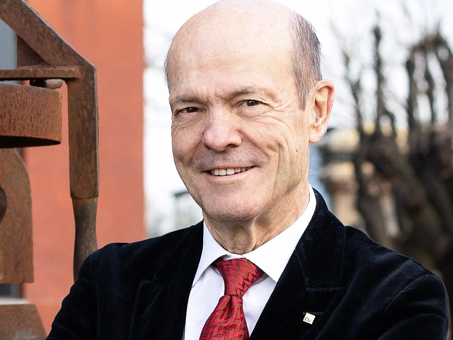 A smiling older man wearing a black jacket, white shirt, and red tie, standing outdoors near an iron railing with trees in the background.