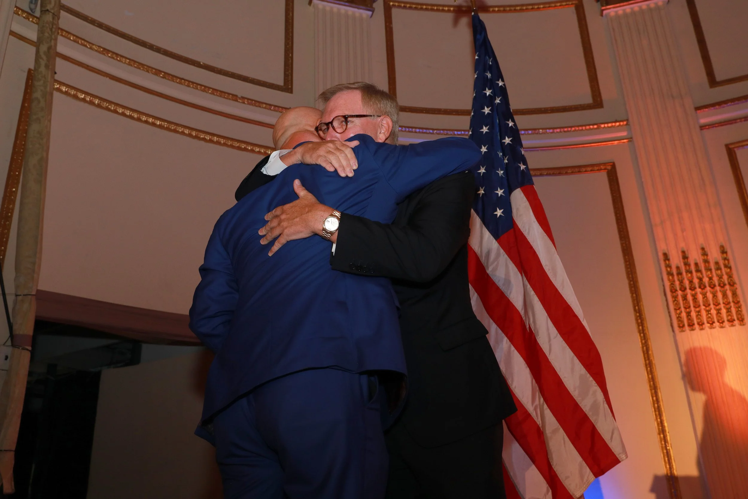 Two men hugging on stage with an American flag behind them in a room with decorative gold trim