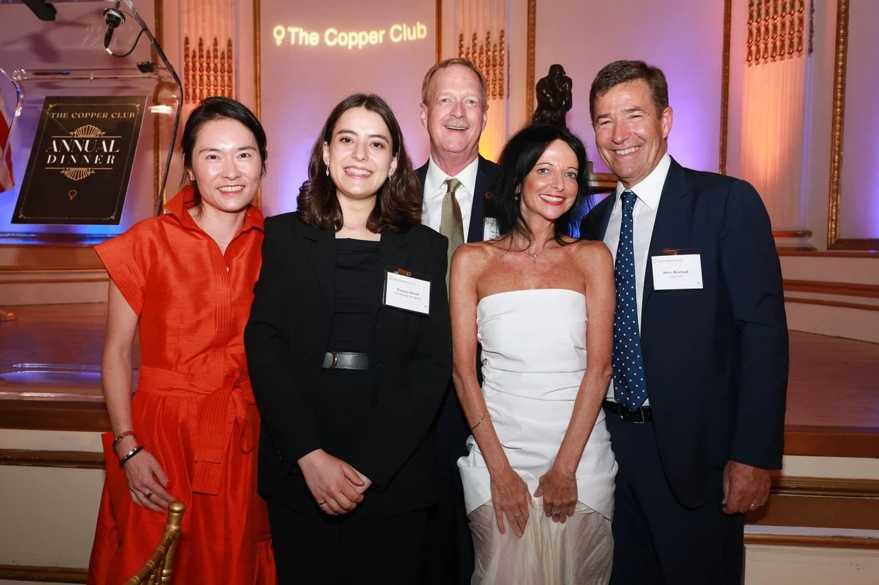 Group of five people posing in formal attire at The Copper Club Annual Dinner event, with a sign and stage in the background.