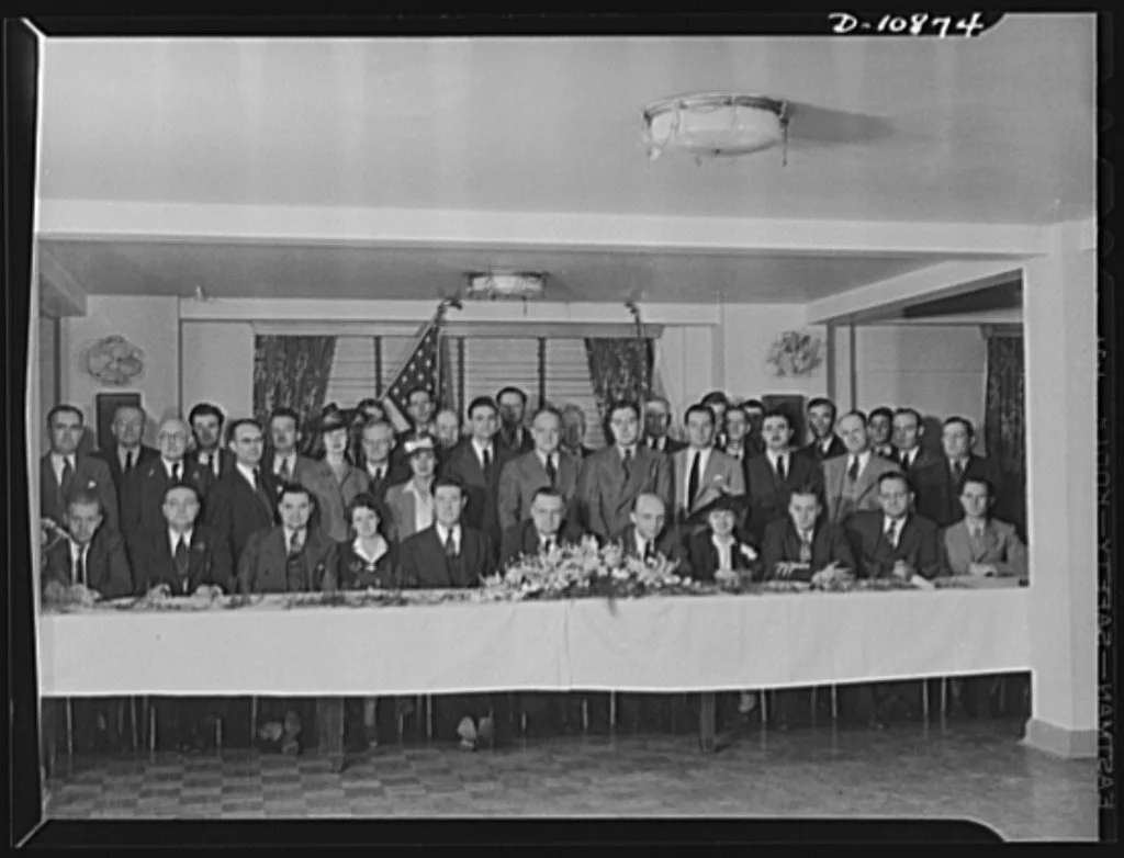A group of men and women in formal attire gathered around a long table in a room with an American flag, curtains, and floral decorations.