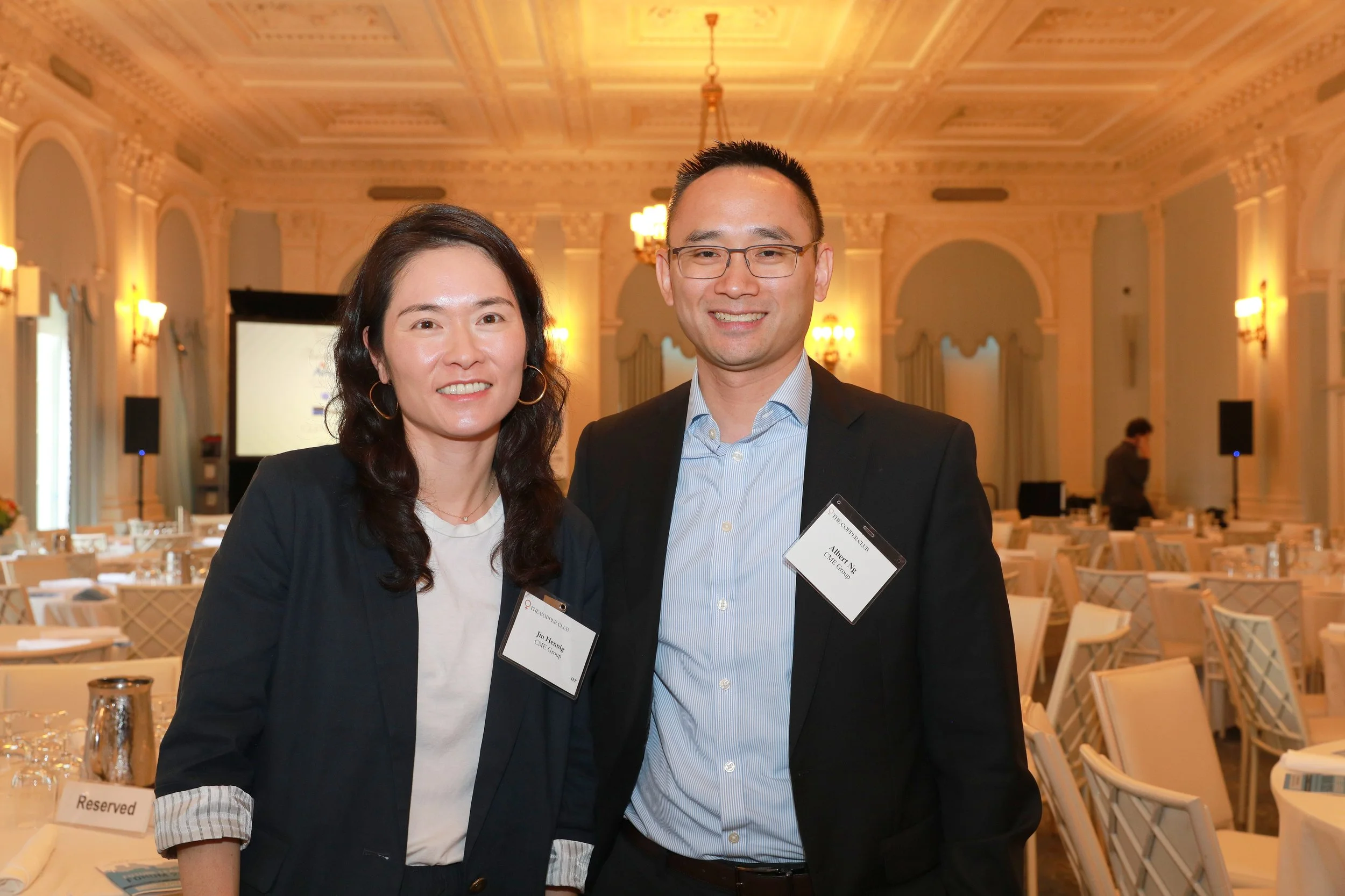 Two people, a woman and a man, standing together at a formal event in an elegant banquet hall. They are wearing name tags and smiling at the camera. The hall has decorated walls, high ceilings, and tables arranged in the background.