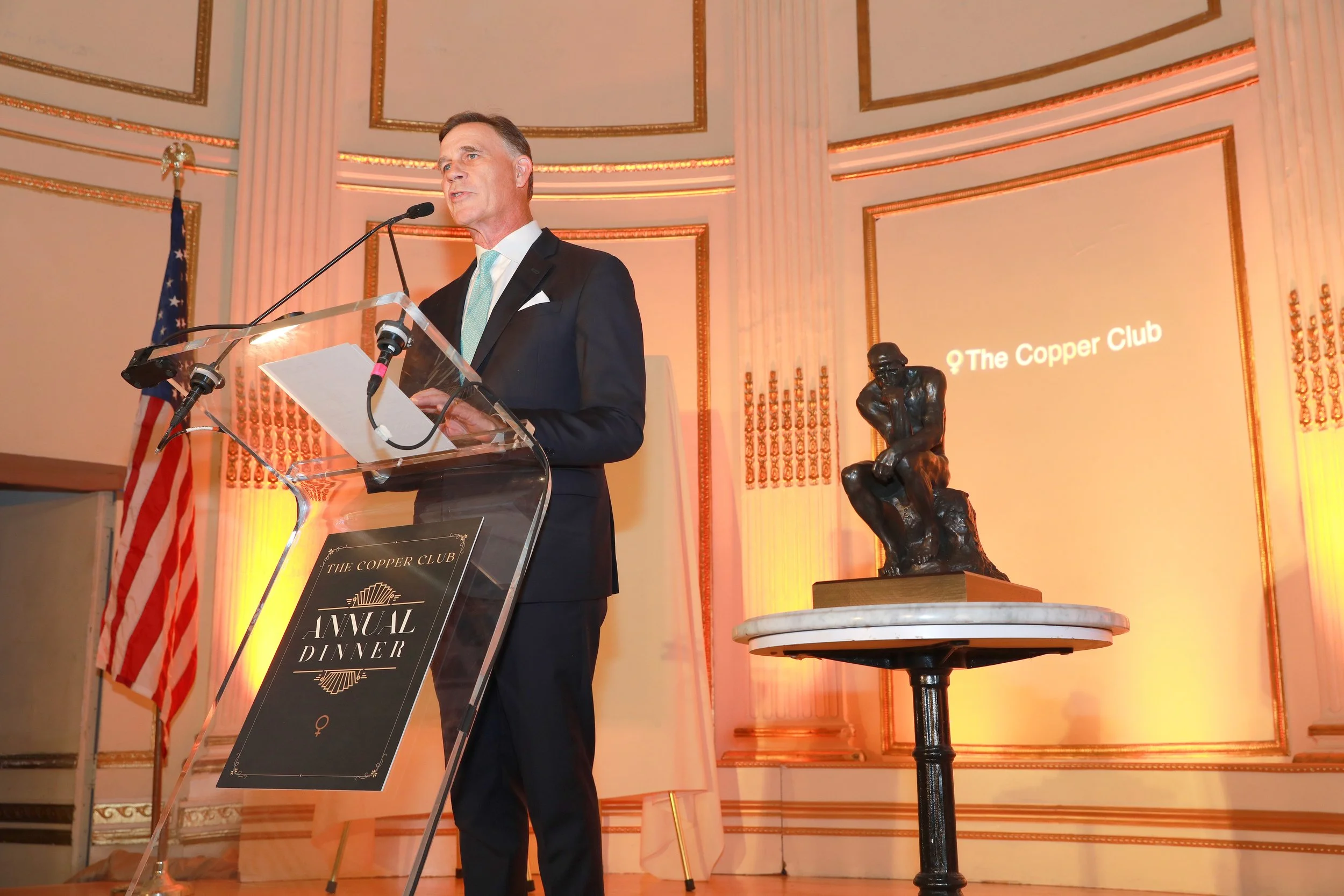 A man in a dark suit and light blue tie speaking at a podium during an event called 'The Copper Club Annual Dinner.' There is a sculpture of a thinker on a table to the right of the speaker and an American flag behind him. The background features ornate gold accents and a large screen with the event name.