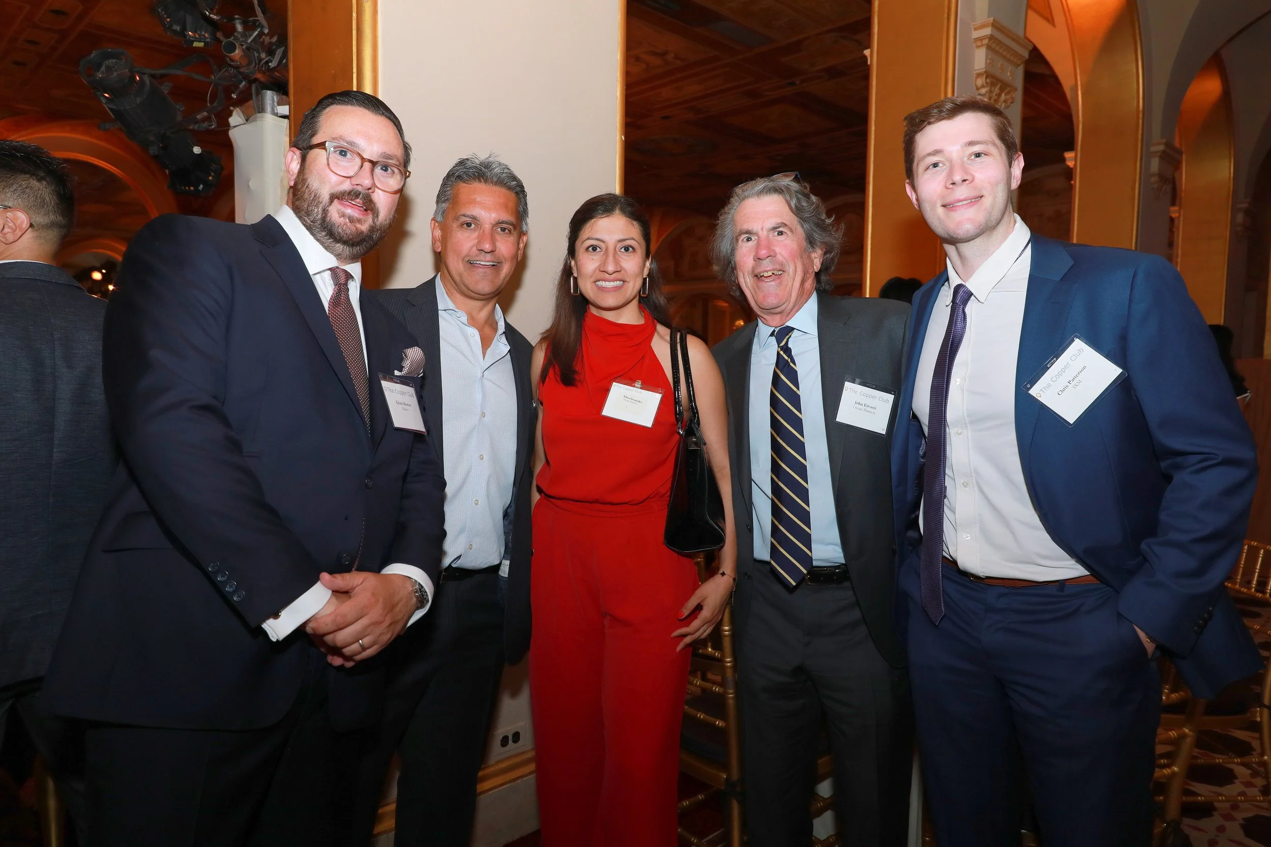 Five people dressed in formal attire standing together at an event with a wooden ceiling and lit arches in the background. They are smiling at the camera, with two men in suits, two women, and one man with gray hair, all wearing name tags.