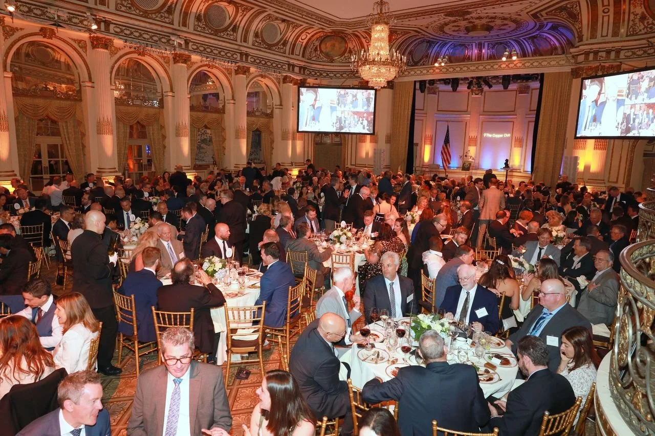 A large formal banquet hall filled with attendees seated at round tables, with some standing and conversing. The room has ornate decor with large windows, chandeliers, and a stage with a screen and American flag.