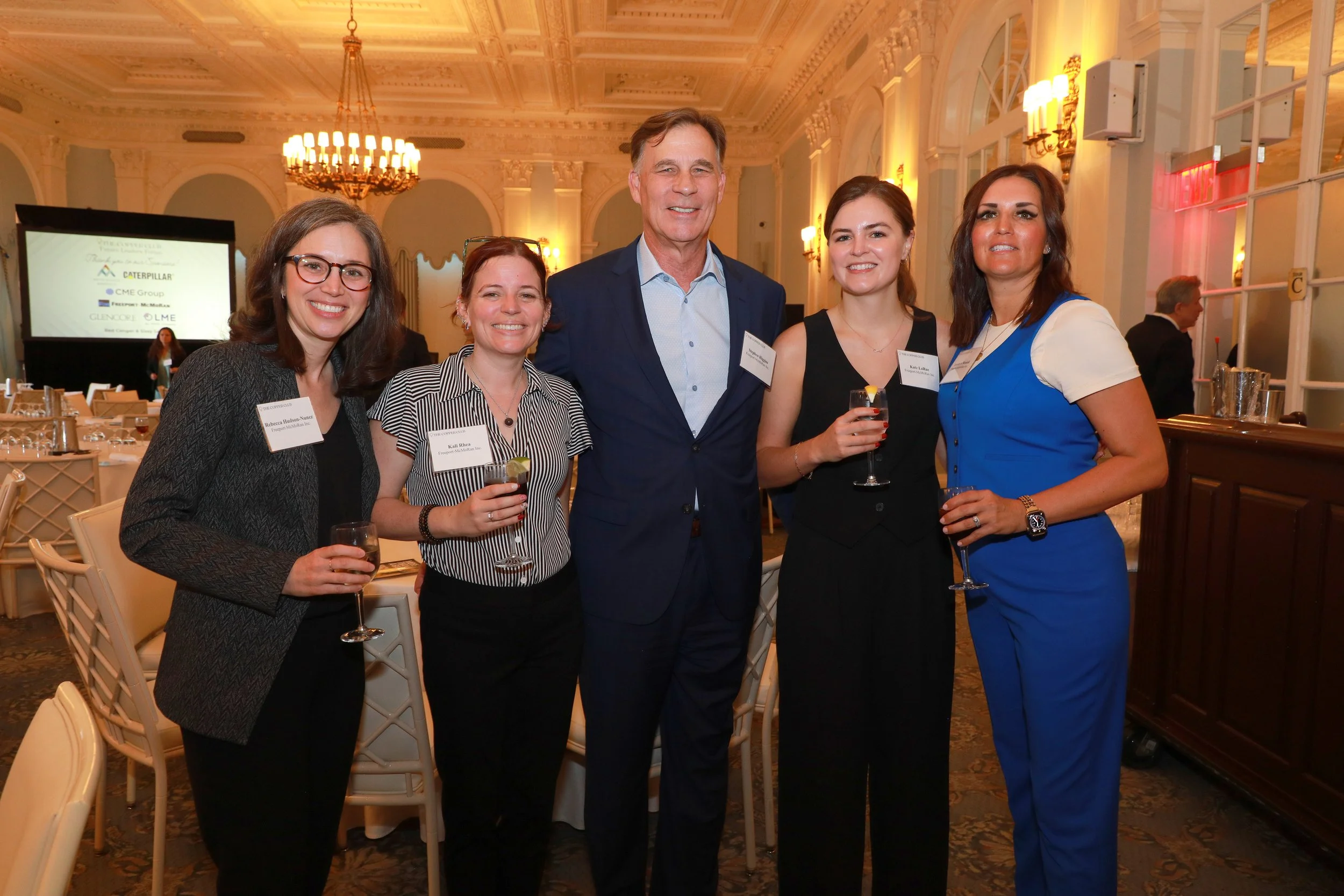 A group of five people at a formal event, smiling and holding drinks, in a luxurious decorated ballroom with chandeliers and large windows.