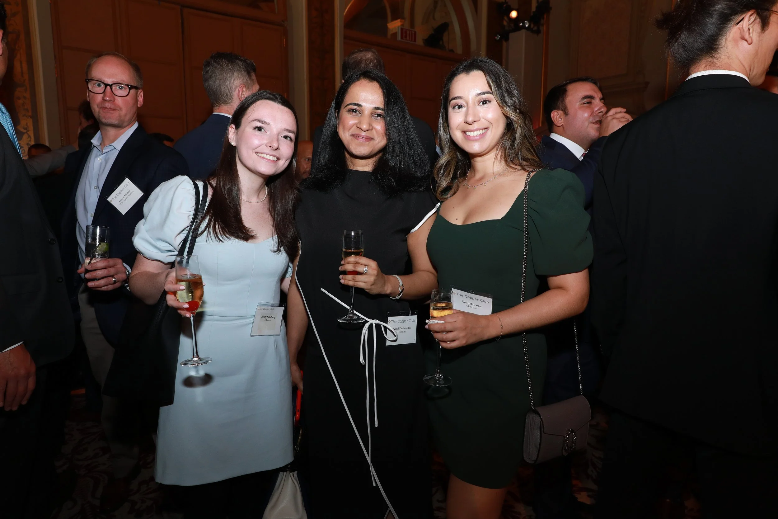 A group of women and men at a formal event, with three women in the foreground holding glasses of champagne, dressed in elegant attire, smiling at the camera.